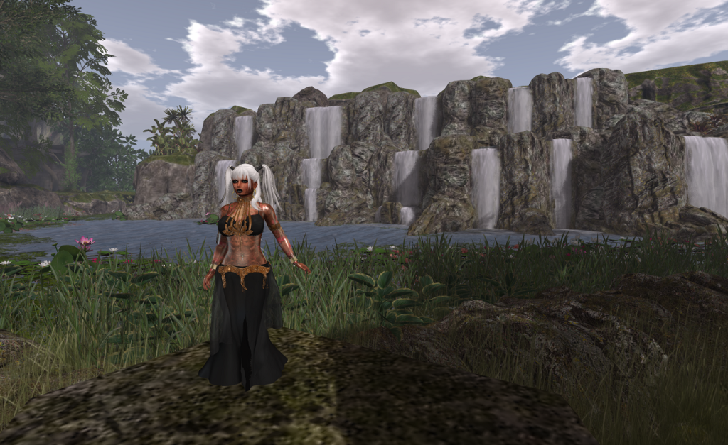A woman with long white hair tied in pigtails stands on a broad, flat stone in the foreground. She is wearing a black and gold, long skirted belly dancer's outfit. Just behind her is a grassy bank before a large lake. In the distance, on the far side of the lake, half a dozen water falls spill into the lake in two step cascades.
